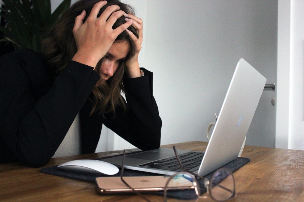 Kobieta w czarnym ubraniu siedząca przy biurku z laptopem, trzymająca głowę w dłoniach, wskazując na oznaki stresu i przeciążenia.