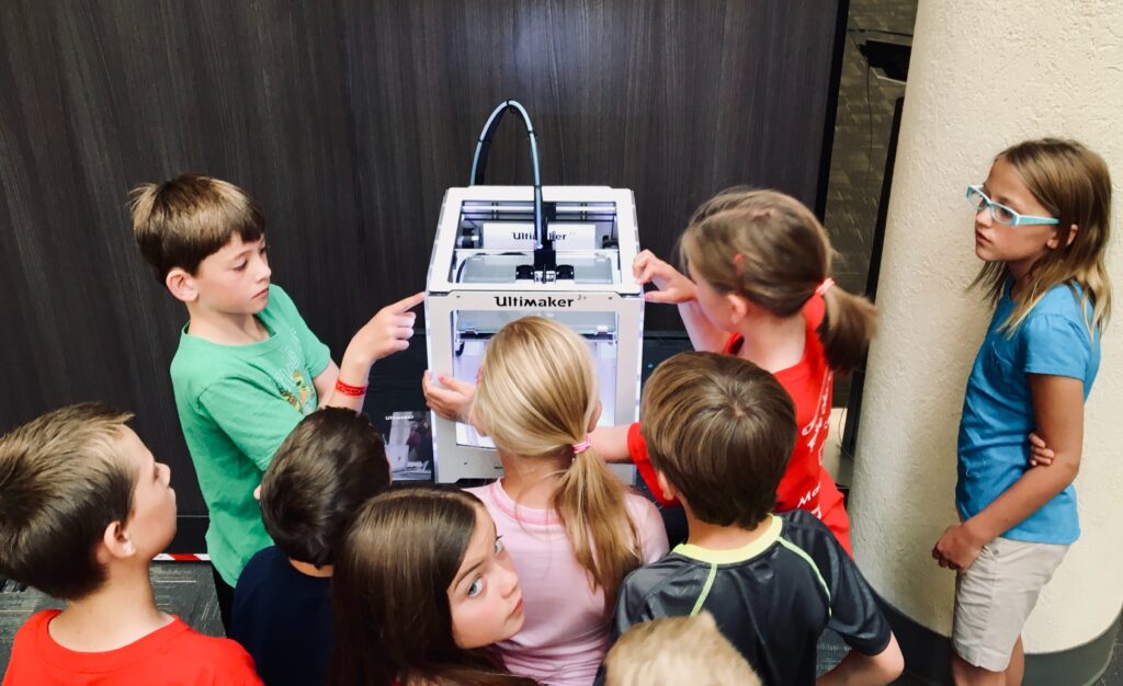 Dzieci w wieku szkolnym skupione na oglądaniu działania drukarki 3D podczas zajęć edukacyjnych, zaintrygowane nowymi technologiami i ich możliwościami.