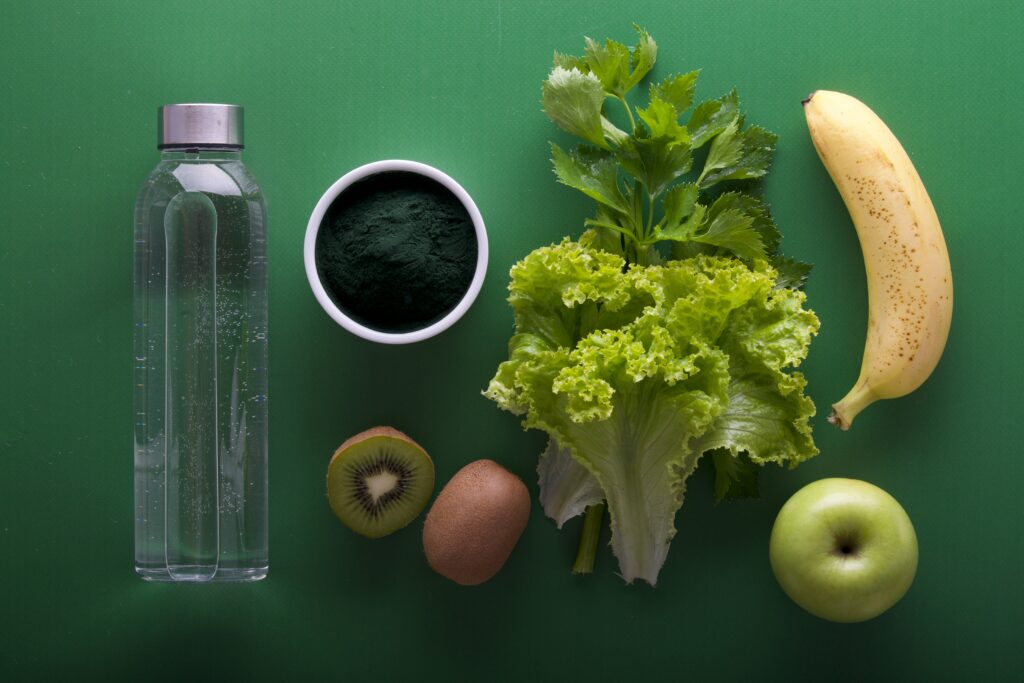 Butelka wody, spirulina w misce, świeży banan, kiwi, jabłko i sałata na zielonym tle, komponują zdrowy i zbilansowany zestaw żywieniowy
