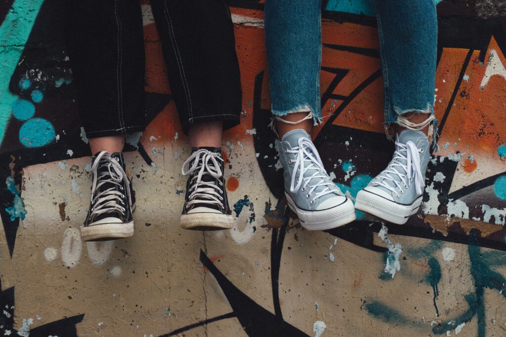Dwie pary nóg w casualowych sneakersach zwisających nad kolorowym graffiti, odzwierciedlające młodzieżowy styl i subkulturę.