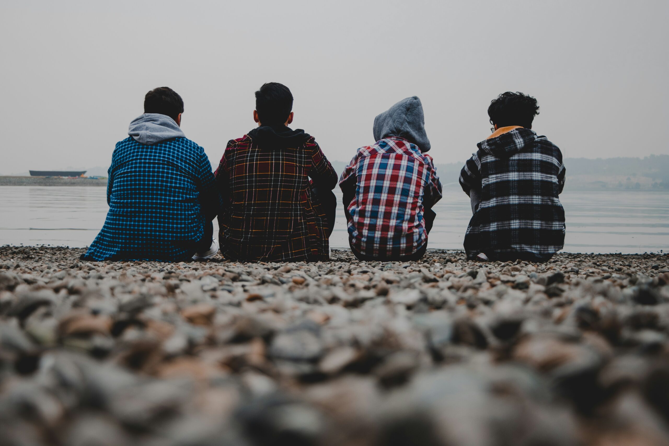 Czterech nastolatków siedzi obok siebie, opierając się plecami o kamienisty brzeg jeziora, co symbolizuje jedność i wsparcie wśród młodzieży.