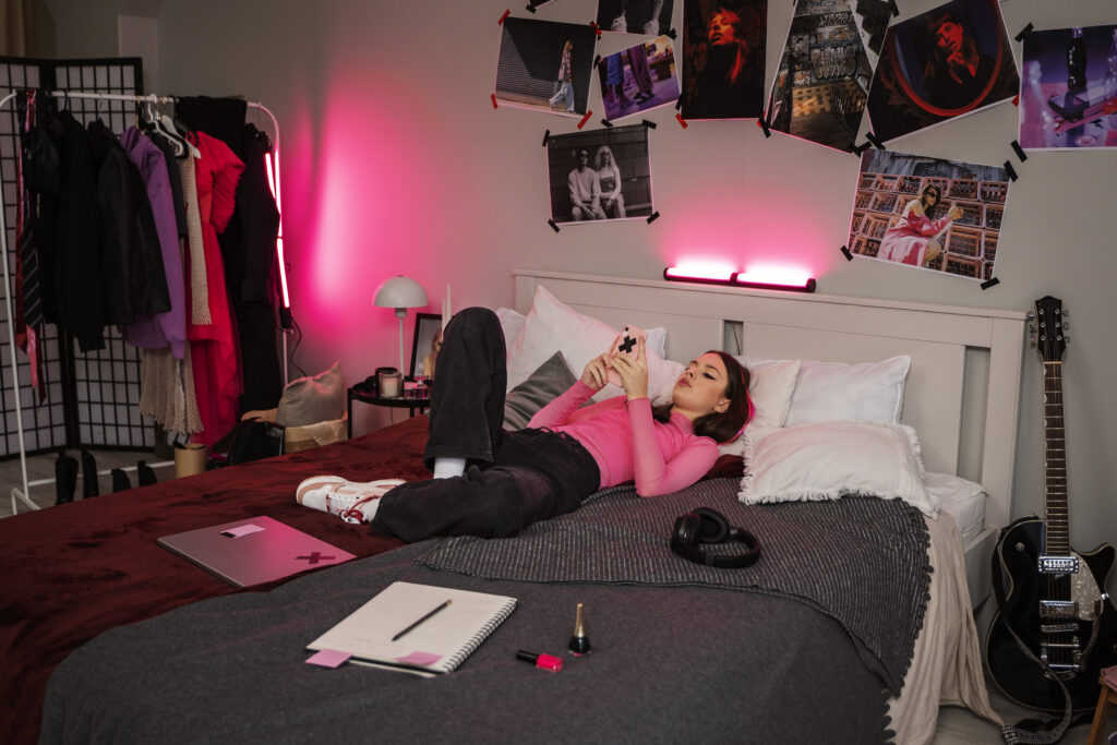 Nastolatka leży na łóżku w swoim pokoju, otoczona personalizowanymi dekoracjami, przeglądając telefon, w tle widoczne są muzyczne i modowe akcenty oraz dynamiczne oświetlenie.