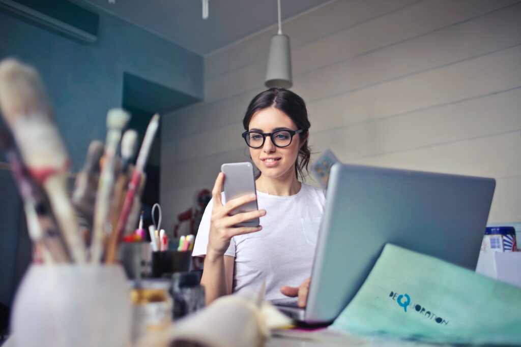 Kreatywna młoda kobieta w okularach, pracująca na laptopie i sprawdzająca swojego smartfona w artystycznie nieuporządkowanym biurze domowym.
