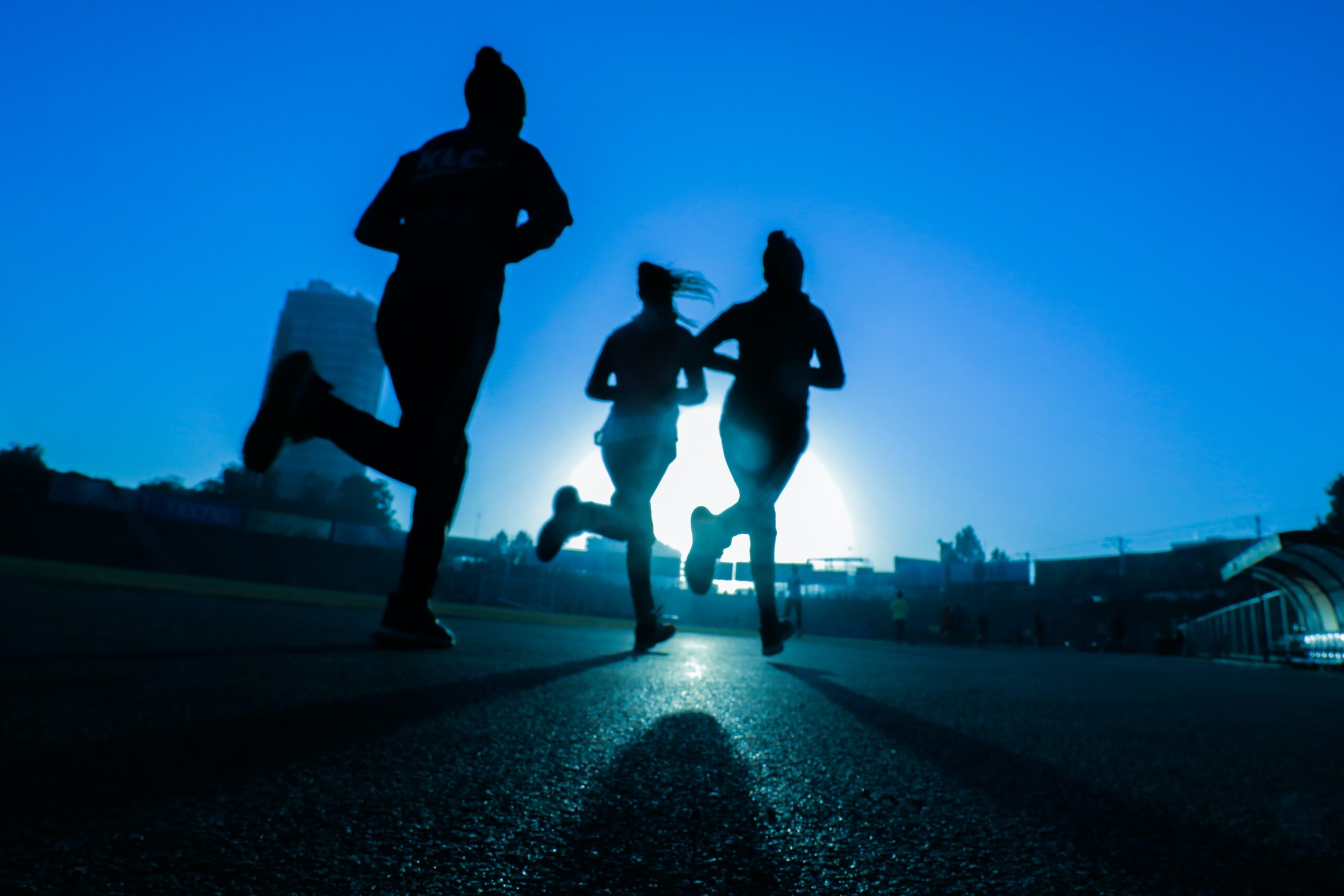 Sylwetki trzech biegaczy biegających po bieżni o świcie, na tle wschodzącego słońca, symbolizujące witalność i zdrowy tryb życia